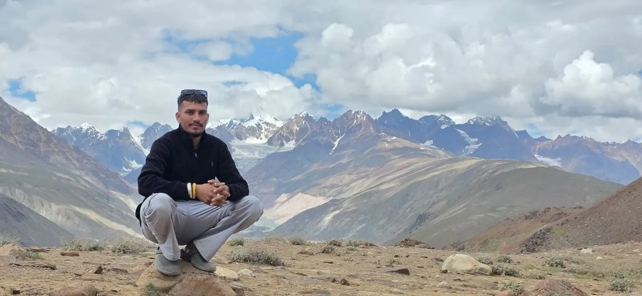 Kulwant crouching on a ridge above Chandratal, snow-capped peaks behind him
